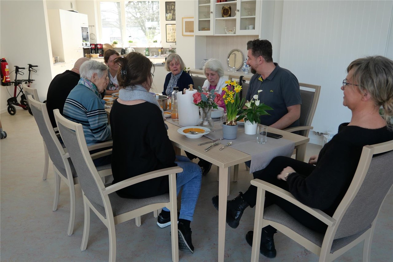 Gruppe von Personen an einem gedeckten Esstisch in einem hellen Raum in der Tagespflege Franziskus. Auf dem Tisch stehen Teller, Besteck, Gläser, Speisen und Blumenarrangements. Im Hintergrund weiße Wände, Schränke, Dekorationen und Küchengeräte.