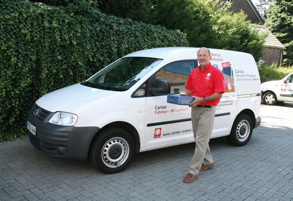 Person in rotem Shirt und beiger Hose steht neben einem weißen Lieferwagen mit der Aufschrift „Caritas Fahrbarer Mittagstisch“ und der Webadresse „www.caritas-krefeld.de“.