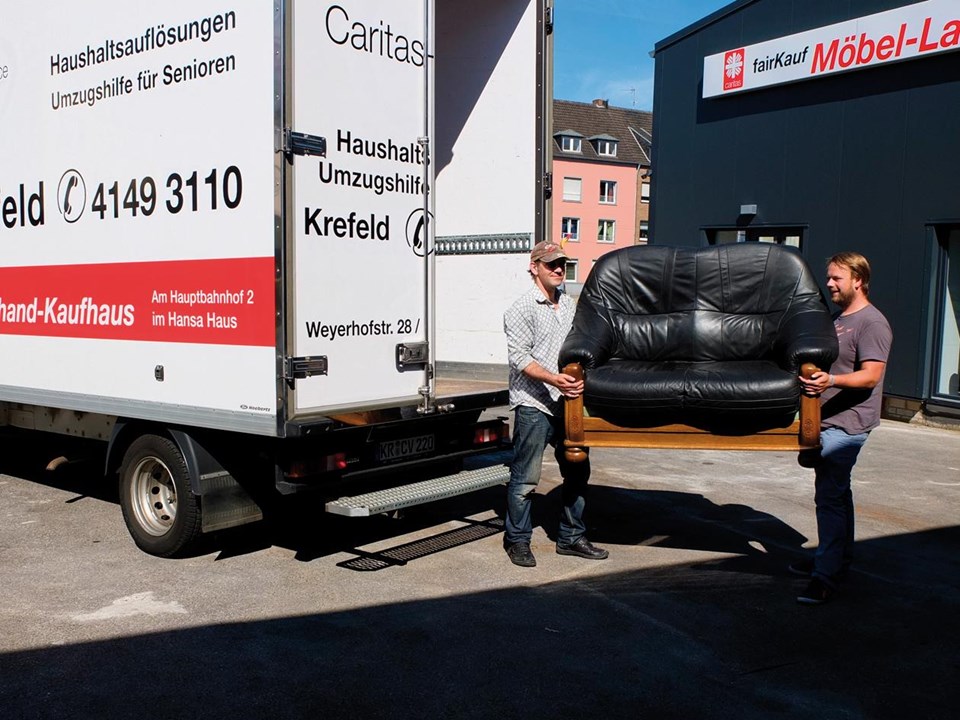 Zwei Personen tragen ein schwarzes Ledersofa zu einem Umzugswagen mit Aufschrift „Haushaltsauflösungen – Umzugshilfe für Senioren – Caritas Krefeld“; im Hintergrund ist ein Gebäude mit dem Schild „fairKauf Möbel-Laden“ zu sehen.