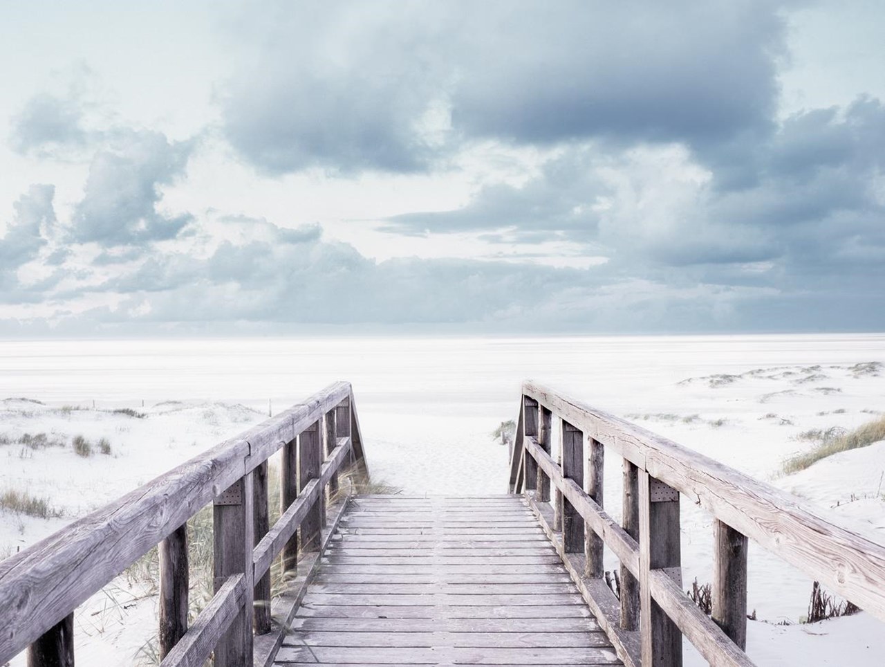 Holzsteg mit seitlichen Geländern führt über sandige Dünen mit spärlicher Vegetation in Richtung Strand. Der Himmel ist von großen, dunklen Wolken bedeckt und verleiht der Szenerie eine ruhige, leicht düstere Stimmung.